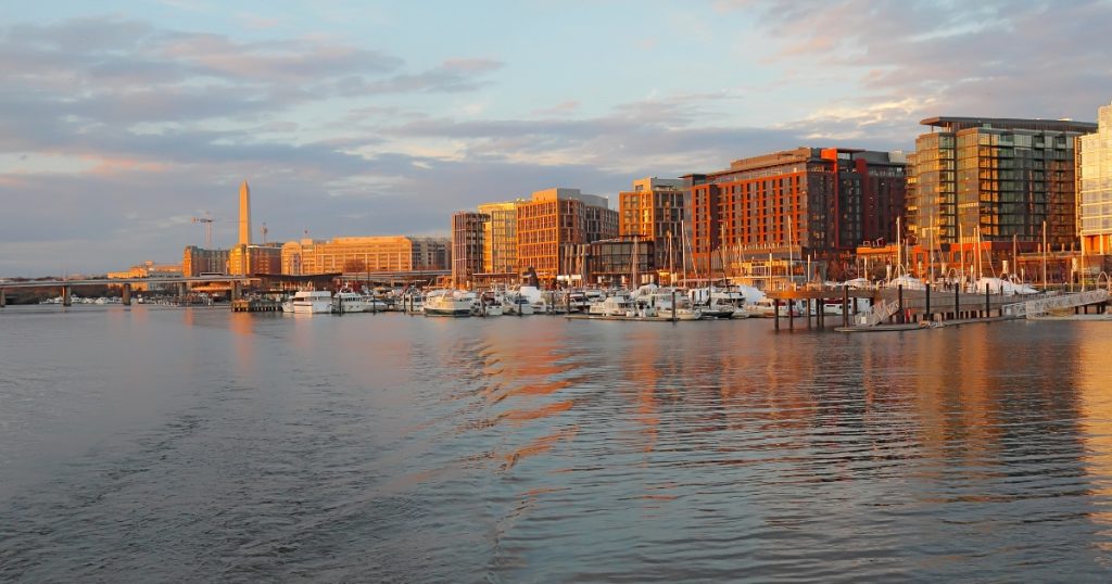The Wharf waterfront district in Washington, D.C., with boats docked in the marina, modern buildings, and the Washington Monument visible in the distance at sunset.