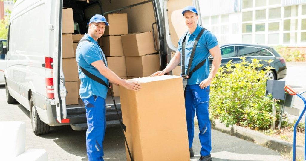 Two professional movers unloading a large cardboard box from a moving truck.